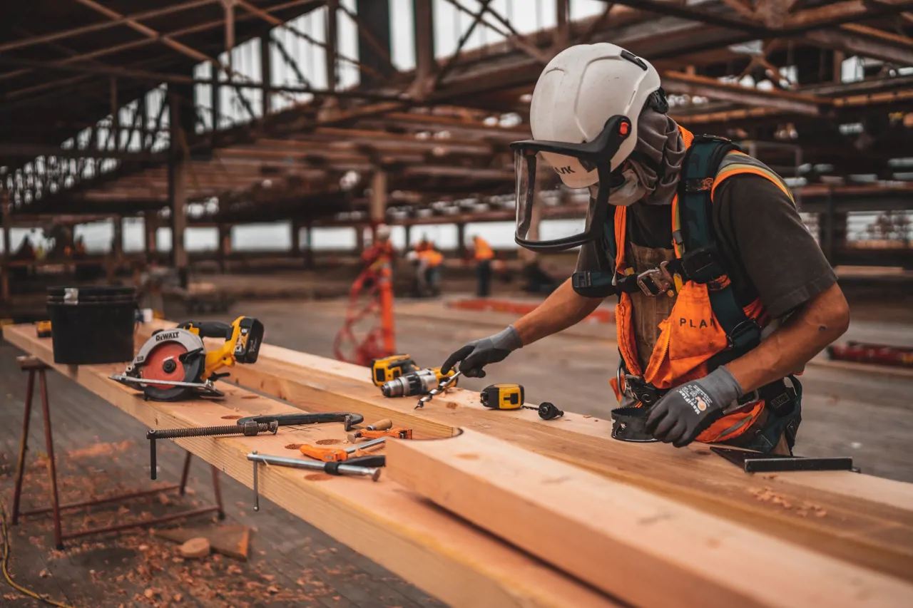 A worker in safety gear uses tools on a wooden beam at a construction site, with various tools scattered around.